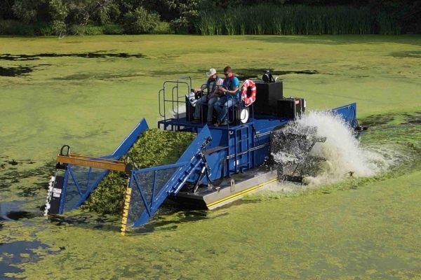 Aquarius Systems HM-420 Midsize Harvester - Eurasian Milfoil and Algae Dark blue weed harvester in lake with surface water covered in green algae and milfoil.